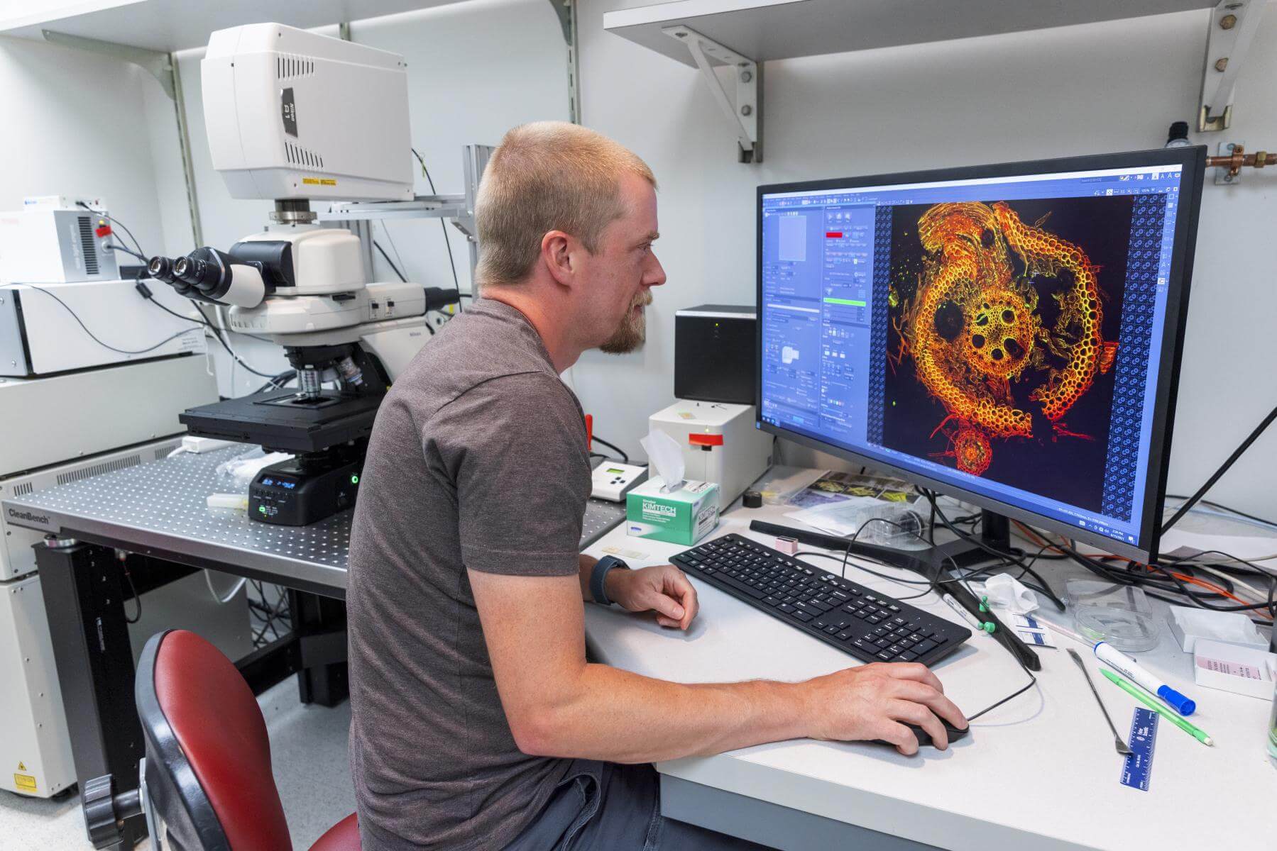 A man sits focusing on an image on a monitor with a microscope over his shoulder.