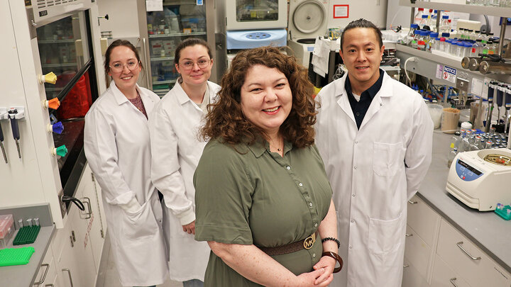 Four University of Nebraska–Lincoln researchers — (from left) Darla Brennan and Madison Broeker, doctoral students in biochemistry; Nicole Buan, professor of biochemistry; and Henry Chen, postdoctoral researcher in biochemistry — pose in a lab. Buan is wearing a green dress and the others are wearing white labcoats.